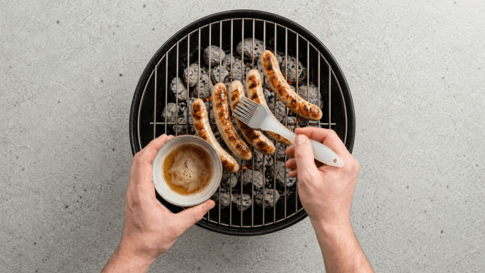 Zwei Hände über einem Holzkohlegrill: mit Pinsel Marinade auf bratende Bratwürste streichen, andere Hand hält eine Tasse.