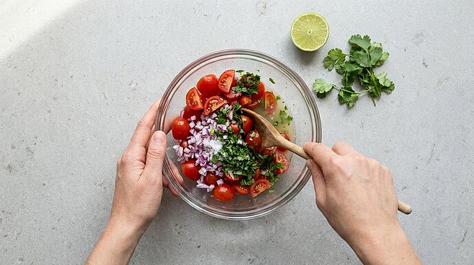Zwei Hände rühren in einer Glasschüssel eine Tomaten‑Koriander‑Salsa mit halbierten Kirschtomaten, fein gehackter roter Zwiebel und gehacktem Koriander; eine Limettenhälfte liegt daneben.