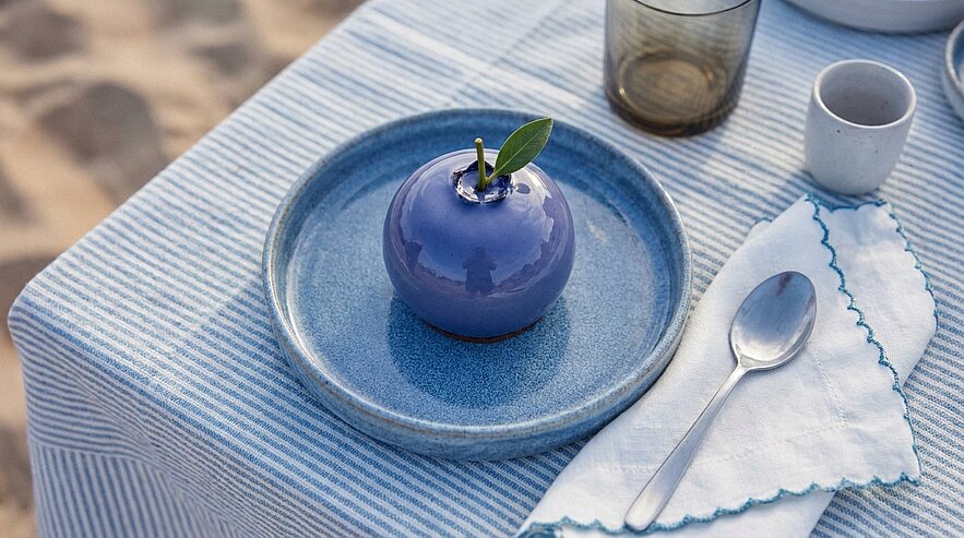 Glänzendes blau-violettes Dessert in Apfelform mit Stiel und Blatt auf blauem Teller; Löffel und weiße Serviette auf gestreifter Tischdecke.