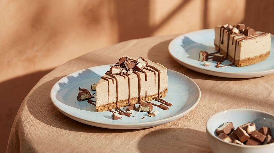 Zwei Stücke Käsekuchen mit Keksboden, Schokoladenglasur und gehackten Schokostücken auf hellblauen Tellern auf einem Tisch im warmen Sonnenlicht.