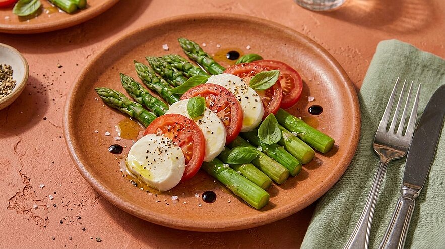 Ein brauner Teller mit einer Portion Tomaten, Mozzarella und grünem Spargel, garniert mit frischen Basilikumblättern. Ein Spritzer Olivenöl und schwarzer Pfeffer verleihen dem Gericht Geschmack. Im Hintergrund sind Textilien und Geschirr sichtbar.