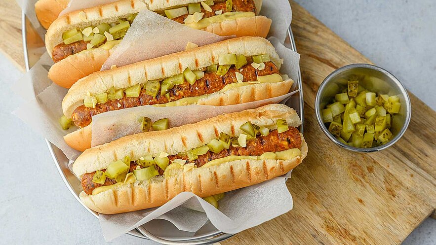 Ein Teller mit mehreren Hotdogs, gefüllt mit einer Wurst und belegt mit gehacktem Gurken. Die Hotdogs sind in Papier gewickelt und liegen in einem metallenen Behälter. Neben dem Teller befindet sich eine kleine Schale mit weiteren gehackten Gurkenstücken.