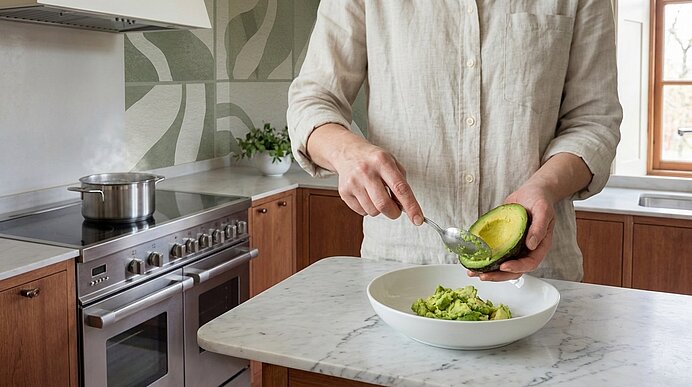 Person löffelt Fruchtfleisch aus einer Avocadohälfte in eine Schüssel auf der Küchenarbeitsplatte.