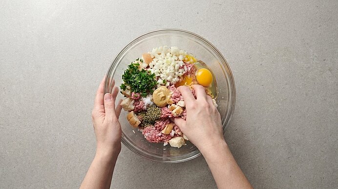 Zwei Hände verkneten Hackfleisch, Ei, Zwiebeln und Kräuter in einer Glasschüssel auf hellgrauer Arbeitsfläche.