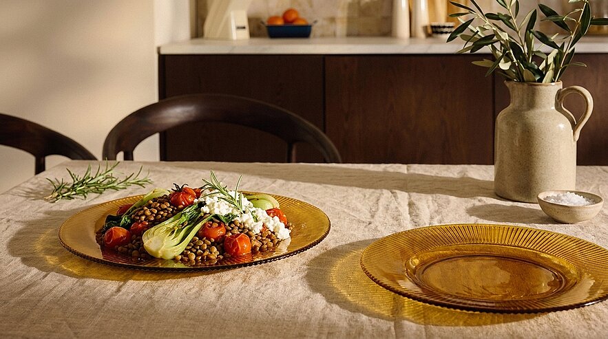 Teller mit Linsensalat, gerösteten Kirschtomaten, Feta und grünen Blättern auf einer gedeckten Leinentischdecke; daneben ein leerer bernsteinfarbener Glasteller und eine Keramikvase mit Zweigen.