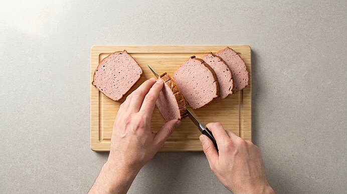 Zwei Hände schneiden mit einem Messer auf einem Holzbrett eine Scheibe Aufschnitt/Wurst; mehrere bereits geschnittene Scheiben liegen daneben.