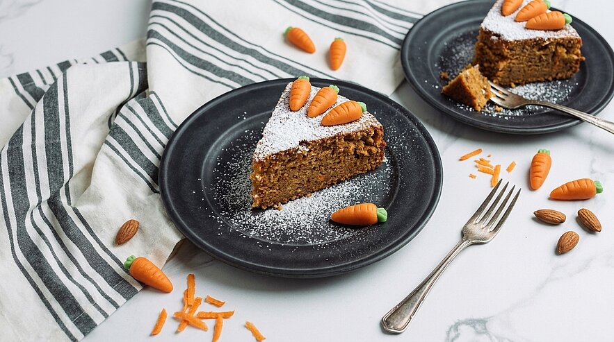 Ein Stück Karottenkuchen auf einem schwarzen Teller, dekoriert mit kleinen Marzipanmöhren und Puderzucker. Daneben liegt eine Gabel. Im Hintergrund ist ein weiterer Teller mit Kuchen zu sehen, umgeben von kleinen Karotten aus Esswaren und einem gestreiften Geschirrtuch.