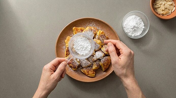 Zwei Hände bestreuen den fertigen Kaiserschmarrn auf einem Teller mithilfe eines Siebs mit Puderzucker.