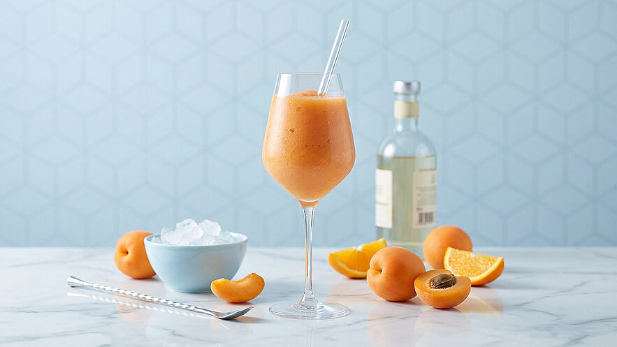 Ein Glas mit einem orangefarbenen Smoothie steht vor einem hellblauen Hintergrund. Daneben liegen frische Aprikosen und eine orangefarbene Scheibe. Eine Schüssel mit Eiswürfeln ist ebenfalls sichtbar, sowie ein langer Löffel.