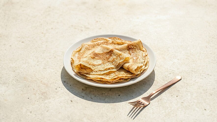 Eine weiße Platte mit mehreren übereinanderliegenden, goldbraunen Crêpes. Sie sind leicht gewellt und haben eine leicht karamellisierte Oberfläche. Neben dem Teller liegt eine roségoldene Gabel auf einer hellen, strukturierten Oberfläche.