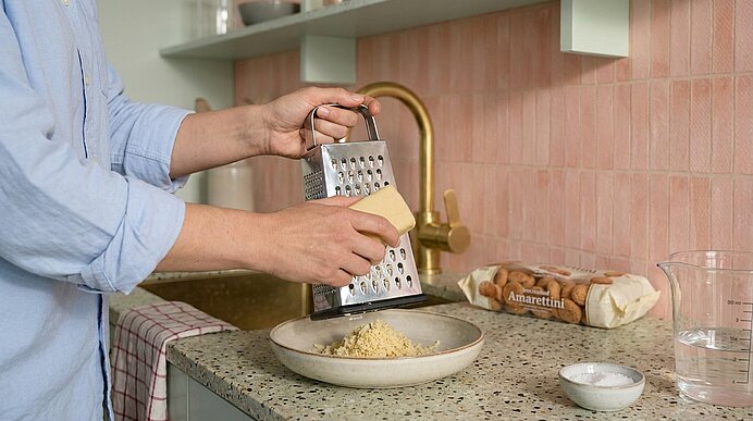 Hände reiben einen Käseblock an einer Kastenreibe über einer Schale, sodass frisch geriebener Käse in die Schale fällt.