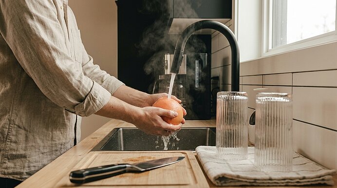 Hände waschen eine Orange unter einem laufenden Wasserhahn in einer Küche; heißer Dampf steigt über dem Spülbecken.