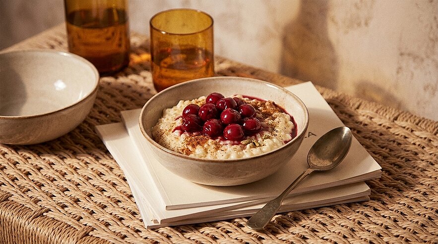 Schale mit cremigem Porridge, bestreut mit Zimt und garniert mit roten Kirschen; ein Löffel liegt daneben.