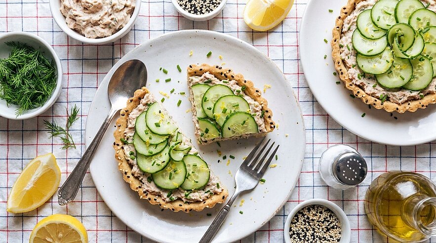 Zwei Gurkentartelettes auf Tellern, eines in Stücke geschnitten; dünne Gurkenscheiben und gehackter Dill auf einer Körnerkruste, dazu Gabel, Löffel, Zitronenspalten und kleine Schälchen mit Aufstrich, Dill, Sesam und Olivenöl.