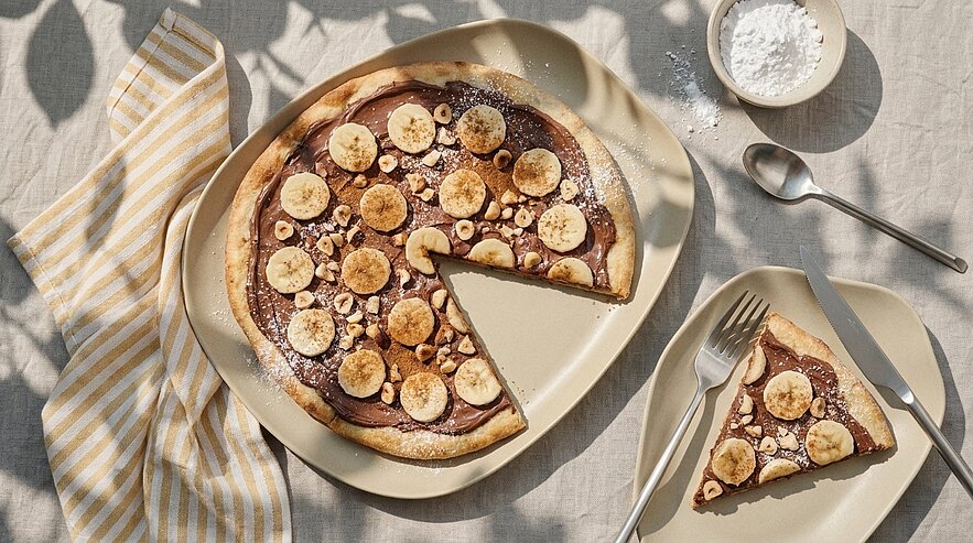 Runder Pfannkuchen mit Schokoladencreme, Bananenscheiben und Puderzucker; ein Stück herausgeschnitten und auf einem Teller serviert.