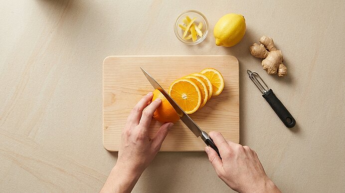 Hände schneiden eine Orange mit einem Küchenmesser auf einem Holz-Schneidebrett; mehrere Orangenscheiben liegen bereits daneben.
