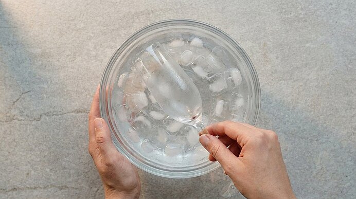 Zwei Hände legen ein Sektglas zum Kühlen in eine Schüssel mit Eiswürfeln auf der hellgrauen Arbeitsfläche.