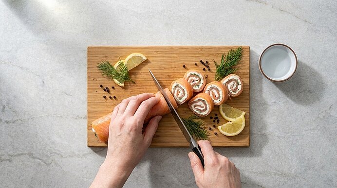 Zwei Hände schneiden eine Lachs-Frischkäse-Rolle auf einem Holzbrett in Scheiben. Garniert mit Zitronenspalten und Dill.