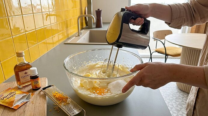 Hände einer Person schlagen mit elektrischem Handmixer Eier und Teig in einer großen Glasschüssel; auf der Arbeitsfläche Vanille, Likör, Zitronenschale und eine Reibe.