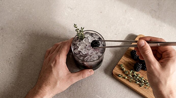 Zwei Hände garnieren ein Glas mit zerstoßenem Eis und dunklem Getränk: eine Brombeere wird mit Zange aufgelegt, daneben Brombeeren und Thymian auf einem Brett.