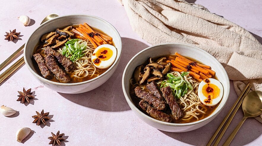 Zwei Schalen mit Ramen-Nudeln, angereichert mit gegrilltem Fleisch, Gemüse und einem halbierten Ei. Die Schalen sind dekorativ angerichtet mit frischen Frühlingszwiebeln und Karottenstreifen, umgeben von Essstäbchen und einem Tuch.