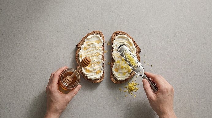 Zwei Hände träufeln Honig und reiben Zitronenschale auf zwei mit Frischkäse bestrichene Brotscheiben.