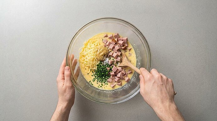 Zwei Hände verrühren Leberkäsewürfel, Käse, Schnittlauch und Eimasse mit einem Holzlöffel in einer Glasschüssel.
