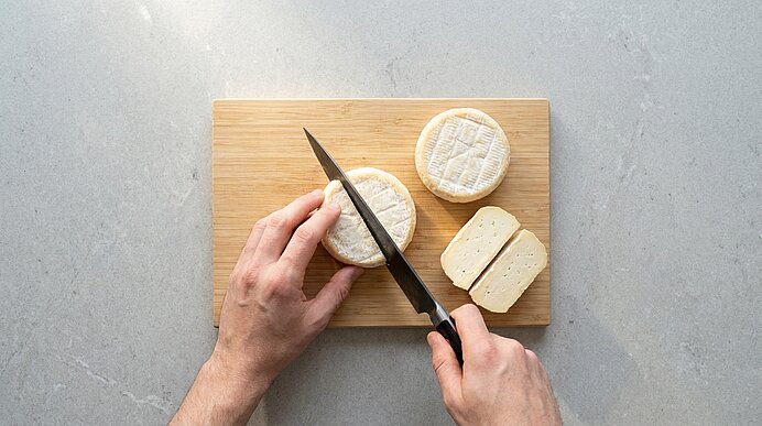Zwei Hände schneiden auf einem Holzbrett einen runden, weichen Käse; daneben liegt ein weiteres rundes Stück und mehrere bereits geschnittene Käsescheiben.