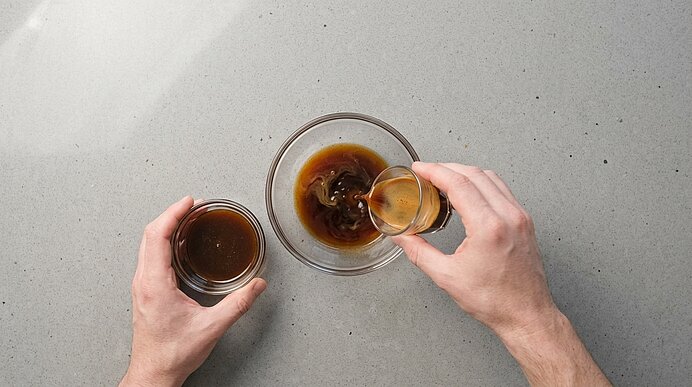 Zwei Hände gießen Espresso aus einem kleinen Glas zu Lebkuchensirup in eine Glasschale auf hellgrauem Naturstein.