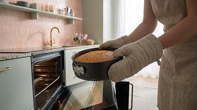 Person in Ofenhandschuhen nimmt eine runde Springform mit frisch gebackenem Kuchen aus dem geöffneten Backofen.