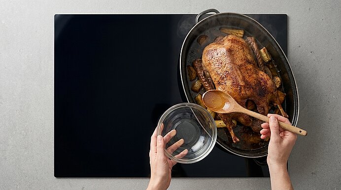Zwei Hände über einer ovalen Bratform: eine hält eine Glasschüssel, die andere bestreicht mit einem Holzlöffel eine goldbraun gebratene, dampfende Ente mit Bratkartoffeln.