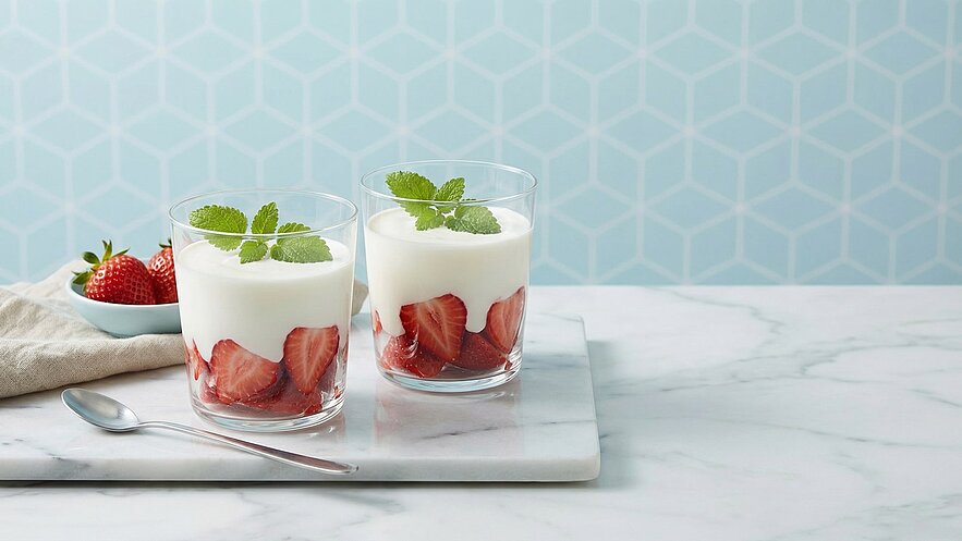 Zwei Gläser mit Joghurt und Erdbeeren stehen auf einem Marmortablett. Die Gläser sind mit geschnittenen Erdbeeren gefüllt und mit Joghurt bedeckt, garniert mit frischen Minzblättern. Im Hintergrund liegen weitere Erdbeeren auf einem kleinen Teller. Der Hintergrund ist hellblau mit geometrischem Muster.