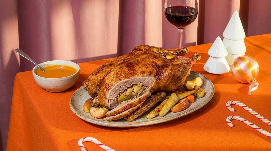 Auf einem festlich gedeckten Tisch: ein goldbraun gebratener, gefüllter Braten auf einer Platte mit Bratkartoffeln und Gemüse, daneben eine Schale Bratensoße und ein Glas Rotwein.