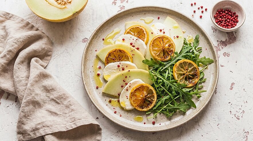 Teller mit aufgeschnittenen Melonenscheiben, Rucola, gerösteten Zitronenscheiben, mit Olivenöl beträufelt und rosa Pfefferkörnern garniert.