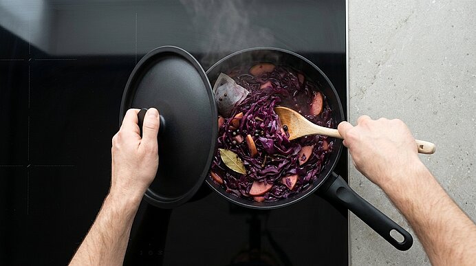 Hände halten einen Deckel und rühren dampfenden Rotkohl mit Apfelscheiben und Gewürzen in einem Topf auf dem Herd.
