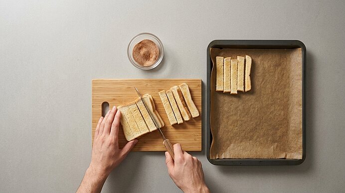 Zwei Hände schneiden entkrustete Toastscheiben auf einem Holzbrett in Stifte. Rechts liegt ein belegtes Backblech.