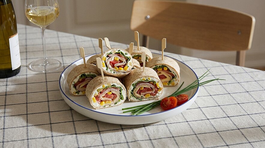 Teller mit aufgeschnittenen Tortilla‑Rolls, gefüllt mit Salat, Schinken, Käse und Gemüse, jeweils mit Holzspieß fixiert, garniert mit Schnittlauch und Kirschtomaten.