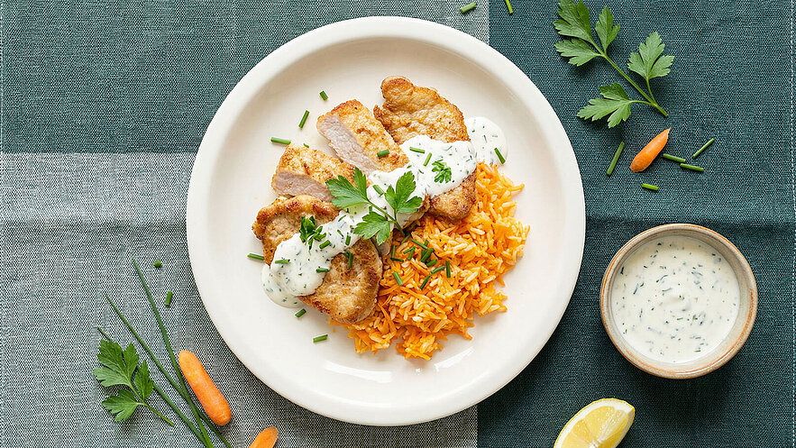 Ein Teller mit gebratenem Hähnchen, garniert mit einer weißen Sauce und frischer Petersilie, liegt auf einem Bett aus orangefarbenem Reis. Daneben gibt es eine Schale mit zusätzlicher Sauce, einen Zitronenschnitz und einige frische Kräuter sowie Karottenstücke.