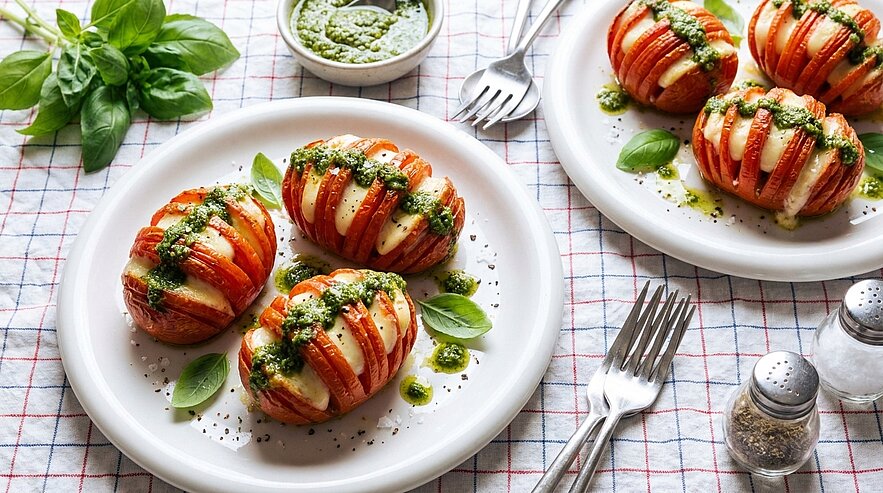 Gebackene Hasselback‑Tomaten: in Scheiben geschnittene Tomaten mit Mozzarellascheiben, beträufelt mit grünem Pesto und garniert mit Basilikum auf weißen Tellern.