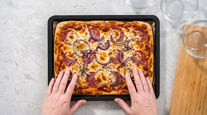 Zwei Hände fassen an ein Backblech mit fertig gebackener Pizza mit Salami, Zwiebeln und Käse.