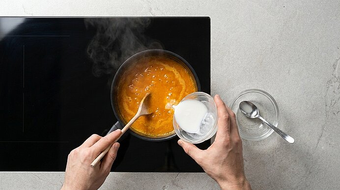 Zwei Hände rühren mit einem Holzlöffel eine orangefarbene Suppe in einem Topf auf dem Herd, während aus einem kleinen Glas Sahne oder Milch dazugegossen wird.