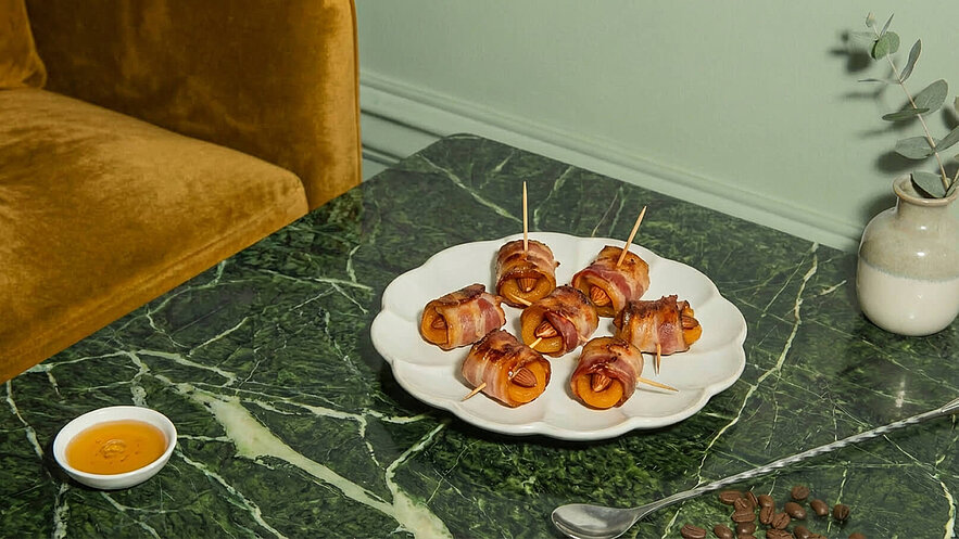 Ein Teller mit kleinen, mit Speck umwickelten Delikatessen, die eine Füllung aus getrockneten Aprikosen und Mandeln enthalten. Daneben befindet sich eine kleine Schale mit einer Sauce. Der Hintergrund zeigt einen marmorierten Tisch und eine sanfte Wandfarbe.