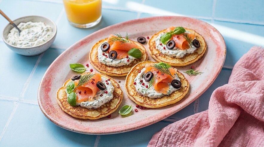 Vier kleine herzhafte Pfannkuchen (Blini) auf einem Teller, jeweils mit Kräuterfrischkäse, Räucherlachs, Olivenringen sowie Dill- und Basilikumgarnitur.
