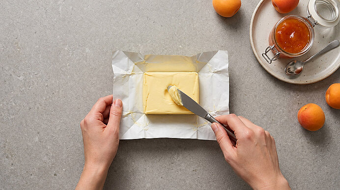 Zwei Hände prüfen die weiche Butter auf hellgrauem Naturstein durch leichten Fingerdruck.