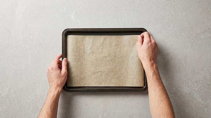 Zwei Hände legen Backpapier auf ein dunkles Backblech auf einer hellgrauen Steinarbeitsplatte.