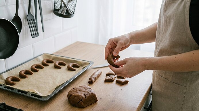 Hände formen aus braunem Teig kleine Gebäckhörnchen auf einer Holzarbeitsfläche; ein Backblech mit bereits geformten Hörnchen liegt daneben, im Hintergrund Küchenutensilien.
