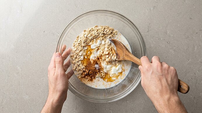 Zwei Hände rühren in einer Glasschüssel Haferflocken, Milch und Joghurt, übergossen mit Honig, mit einem Holzlöffel.