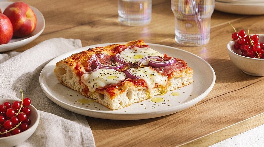 Zwei dicke Stücke luftiger Pizza mit Tomatensauce, geschmolzenem Mozzarella, roten Zwiebelringen und Pfefferoni, leicht mit Olivenöl und Kräutern beträufelt auf einem weißen Teller.