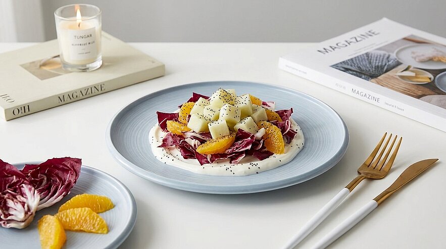 Ein leichtes Salatrezept auf einem blauen Teller, bestehend aus roten Salatblättern, gelben Orangenstücken und weißen Gemüsestücken. Daneben liegt ein kleiner Teller mit weiteren Orangen und eine Kerze brennt im Hintergrund. Auf einem Tisch liegen auch zwei Magazine.