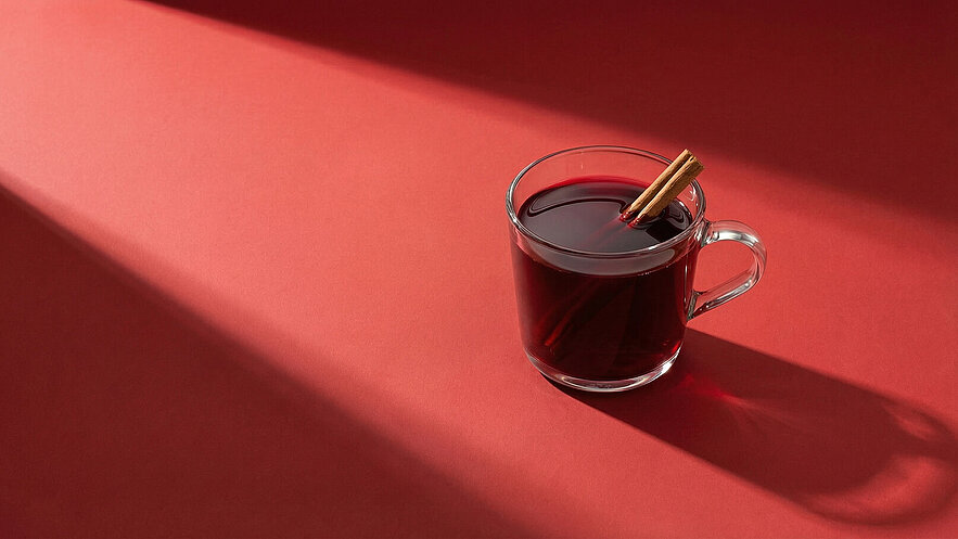Ein Glas mit rotem Getränk, wahrscheinlich Glühwein, steht auf einem roten Untergrund. Zwei Zimtstangen stecken im Getränk, das in warmem Licht beleuchtet wird.